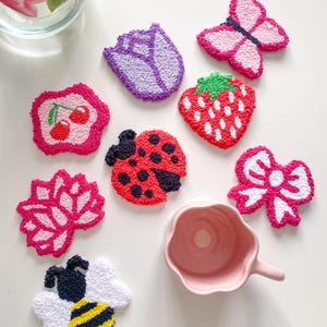 Peut inclure: Divers patchs brodés colorés en forme de papillon, fraise, coccinelle, nœud, abeille et fleur. Une tasse rose est également présente. Les patchs sont disposés sur une surface blanche.