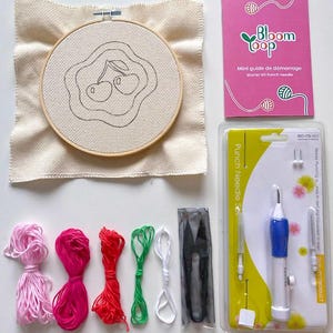 以下が含まれることがあります： 桜のデザインが施されたキャンバスが入った木製フレームの刺繍キット。キットには、ピンク、赤、緑、白の刺繍糸、ハサミ、パンチニードル、そして「Bloom Loop」と書かれたピンク色の小冊子が含まれています。