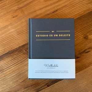 May include: A dark gray journal with gold lettering that reads "MI ESTUDIO ES UN DELEITE". The journal is wrapped in a white label with the text "Worthwhile JOURNALS" and a description in Spanish.