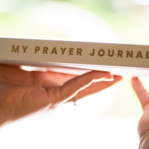 Puede incluir: Un diario blanco con las palabras "MY PRAYER JOURNAL" impresas en letras doradas.
