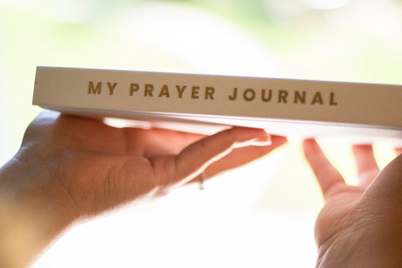 Puede incluir: Un diario blanco con las palabras "MY PRAYER JOURNAL" impresas en letras doradas.