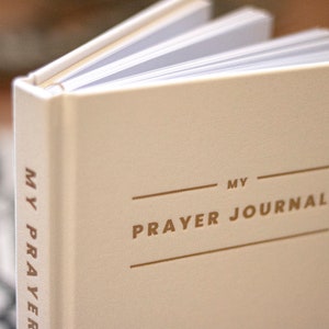 Puede incluir: Un diario de oraci&oacute;n blanco con letras doradas que dicen "MY PRAYER JOURNAL".