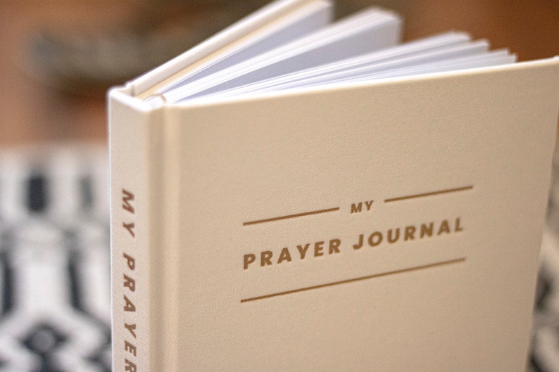 Puede incluir: Un diario de oraci&oacute;n blanco con letras doradas que dicen "MY PRAYER JOURNAL".