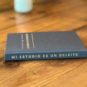 May include: A dark blue notebook with the text "MI ESTUDIO ES UN DELEITE" in gold lettering on the spine.