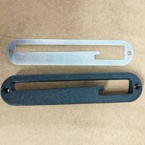 Könnte beinhalten: Zwei Gitarren-Tonabnehmerabdeckungen, eine silberne und eine schwarze. Die silberne Abdeckung ist aus Metall und die schwarze Abdeckung ist aus Kunststoff.