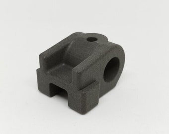 May include: A dark gray, matte-textured mechanical component featuring a U-shaped channel, a central circular opening on the side, and a small, singular hole on the top surface. The object has a blocky, industrial design with defined edges.