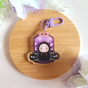 May include: A clear acrylic keychain featuring a purple and white design with a cartoon character and the text "NO FACE". The keychain has a purple clasp and a decorative purple top.