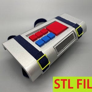 May include: A silver 3D printed prop with a red and blue button panel, a black mesh speaker grille, and two black straps with yellow buckles. STL FILES is printed in red letters on a green background.