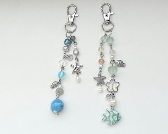 May include: Two silver keychains with blue and green glass beads, starfish, and fish charms. The keychains are on a white background.