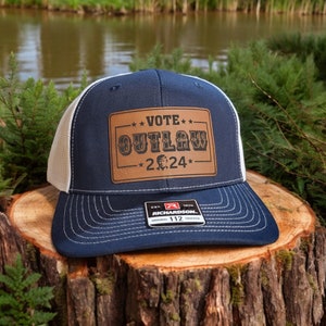 May include: A navy blue and white trucker hat with a brown leather patch that says "***VOTE*** OUTLAW -*224*-". The hat has a white mesh back and a Richardson logo on the side.