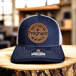 May include: A navy blue and white trucker hat with a brown leather patch that says "Take it easy on Pregnant Me My Wife Is".