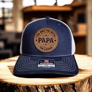 May include: A navy blue and white trucker hat with a brown leather patch that reads "The Man The Myth *Papa* The Legend". The hat is adjustable and has a Richardson logo.