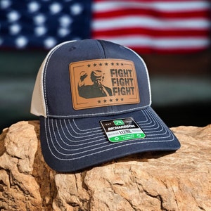 May include: A navy blue trucker hat with a white mesh back. The front of the hat features a brown leather patch with a silhouette of a person with the text "FIGHT FIGHT FIGHT" in white. The hat also has a green and white label with the text "RICHARDSON 112RE RECYCLED TRUCKER FABRIC"