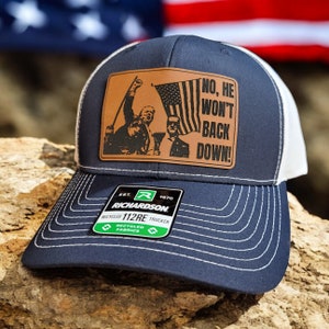May include: A navy blue and white trucker hat with a brown leather patch featuring a political image and the text "NO, HE WON'T BACK DOWN!" The patch is attached to the front of the hat. The hat has a white mesh back and a Richardson logo on the side.