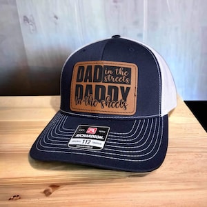Puede incluir: Una gorra de camionero azul marino y blanca con un parche de cuero marrón que dice "Dad in the streets Daddy in the sheets". La gorra es ajustable y tiene un logotipo de Richardson en la parte delantera.