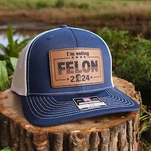 Op de afbeelding: Een blauwe en witte trucker-pet met een bruin leren patch waarop staat "I'm voting FELON *2024*" met een kleine Amerikaanse vlag en sterren op de patch.