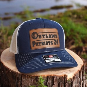 May include: A navy blue and white trucker hat with a brown leather patch that says "Outlaws Patriots '24". The patch features a small image of a person's face inside a circle. The hat has a Richardson 112 label.