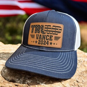 May include: A navy blue and white trucker hat with a brown leather patch that says "TRUMP VANCE 2024" with a small American flag design.