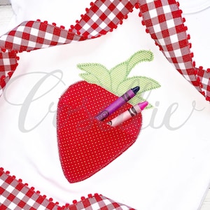 May include: A white t-shirt with a red and white gingham ribbon trim. The shirt features an embroidered red strawberry with green leaves and white polka dots. Two crayons are placed inside the strawberry.