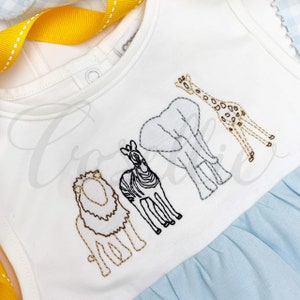 May include: White baby onesie with a blue ruffle at the bottom. The onesie has an embroidered design of a lion, zebra, elephant, and giraffe.