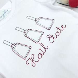 May include: White baby onesie with three gray jingle bells and the text "Hail State" embroidered in maroon thread.
