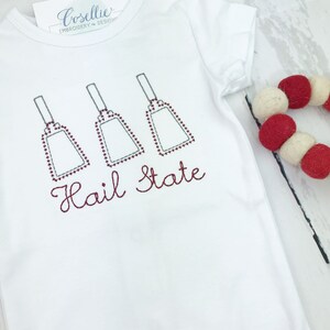 May include: White baby onesie with red embroidered bells and the text "Hail State".