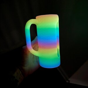 May include: A cylindrical mug with a side handle, featuring a vibrant, glowing horizontal gradient pattern that transitions through shades of yellow, green, blue, and purple. The surface appears to emit light, creating a luminous effect.