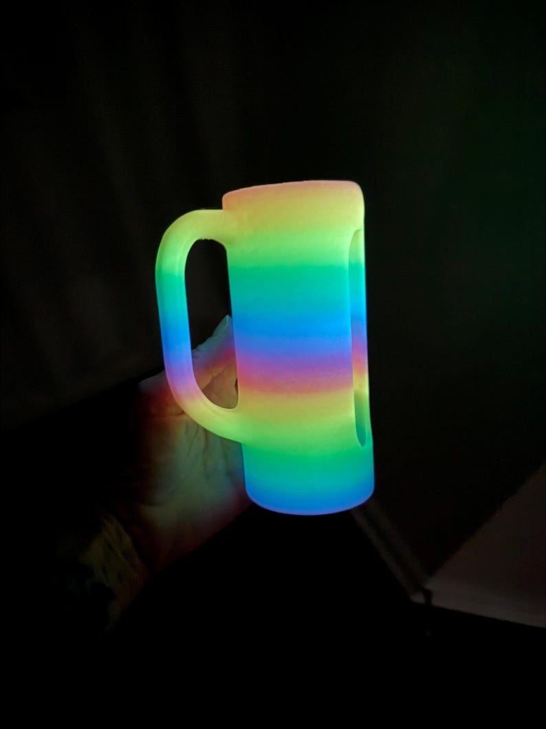 May include: A cylindrical mug with a side handle, featuring a vibrant, glowing horizontal gradient pattern that transitions through shades of yellow, green, blue, and purple. The surface appears to emit light, creating a luminous effect.