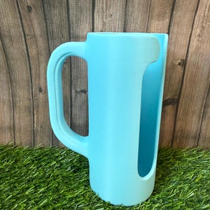 May include: A light blue cylindrical cup holder with a large, curved side handle. The object features a vertical cutout opening along the front side, revealing the interior space. The surface has a matte finish with visible 3D printed texture.