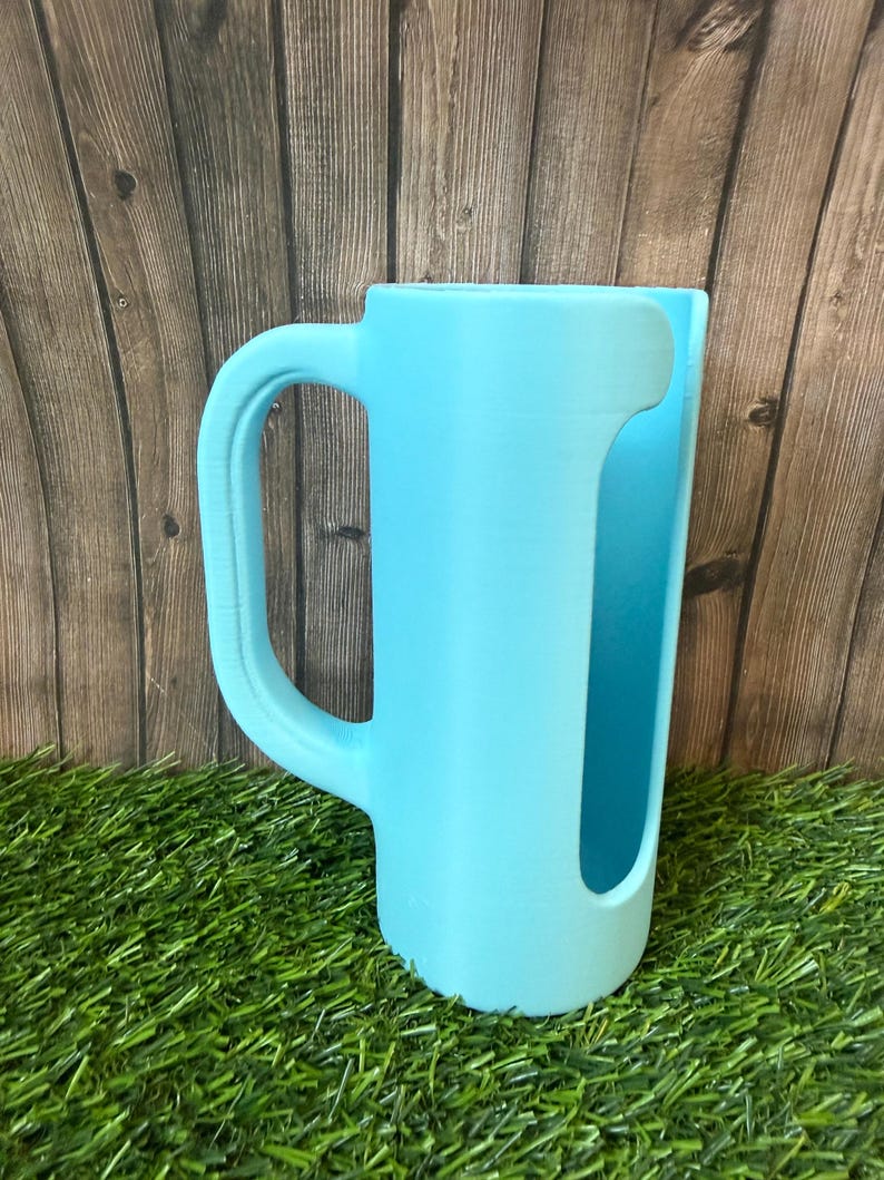 May include: A light blue cylindrical cup holder with a large, curved side handle. The object features a vertical cutout opening along the front side, revealing the interior space. The surface has a matte finish with visible 3D printed texture.