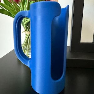 May include: A bright blue cylindrical pitcher with a large, curved handle on the side and a wide, vertical cutout opening extending from the top rim down the front of the vessel, featuring a smooth, matte finish and a hollow interior structure.