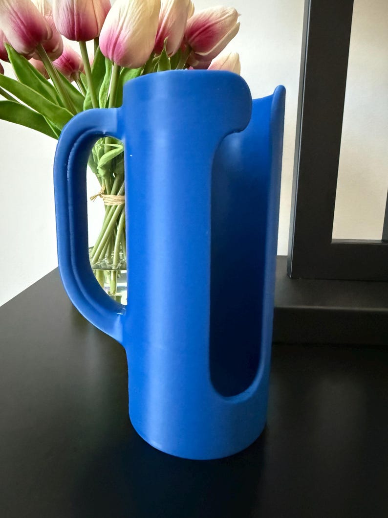 May include: A bright blue cylindrical pitcher with a large, curved handle on the side and a wide, vertical cutout opening extending from the top rim down the front of the vessel, featuring a smooth, matte finish and a hollow interior structure.