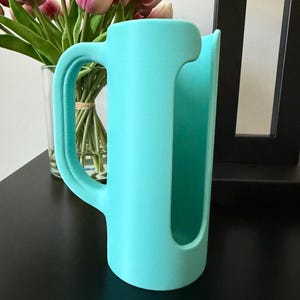 May include: A matte turquoise cylindrical holder featuring a large, curved side handle and a vertical cutout opening along the front. The object has a smooth, solid-colored surface and a hollow interior, designed with a wide base for stability.