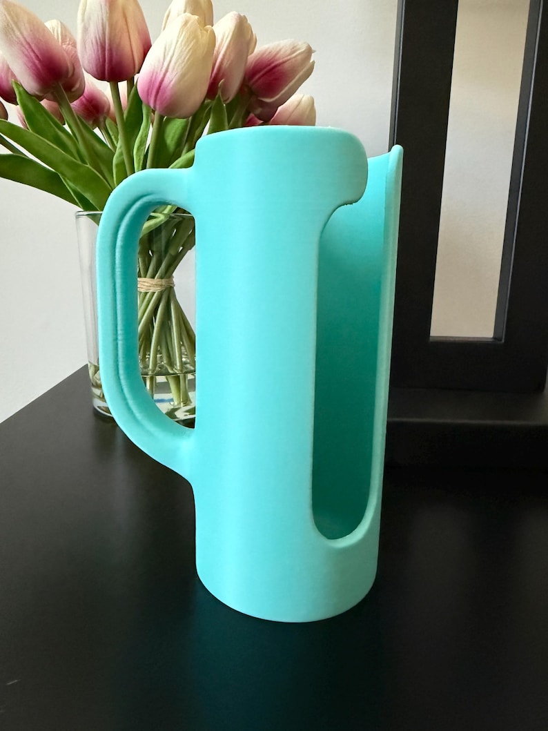 May include: A matte turquoise cylindrical holder featuring a large, curved side handle and a vertical cutout opening along the front. The object has a smooth, solid-colored surface and a hollow interior, designed with a wide base for stability.