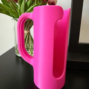 May include: A bright neon pink cylindrical holder featuring a vertical cutout opening and a curved side handle. The object has a smooth, matte finish and a hollow interior, designed with a sturdy base to stand upright on a flat surface.
