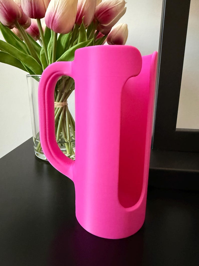 May include: A bright neon pink cylindrical holder featuring a vertical cutout opening and a curved side handle. The object has a smooth, matte finish and a hollow interior, designed with a sturdy base to stand upright on a flat surface.