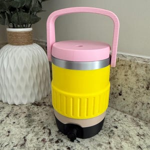 May include: A cylindrical beverage cooler featuring a bright yellow body with a textured middle band, a light pink lid with a matching pink handle, a beige lower section, and a black base equipped with a small black spigot for dispensing liquids.