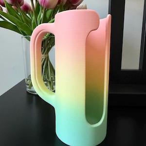 May include: A cylindrical cup holder with a handle, featuring a smooth matte finish and a vertical gradient color scheme that transitions from a soft pink at the top to a bright yellow in the middle and a light mint green at the base.