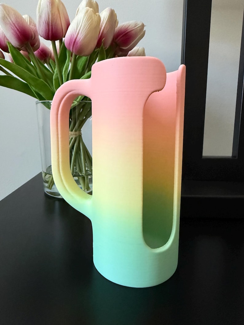 May include: A cylindrical cup holder with a handle, featuring a smooth matte finish and a vertical gradient color scheme that transitions from a soft pink at the top to a bright yellow in the middle and a light mint green at the base.