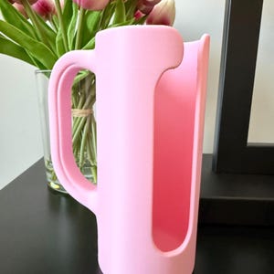May include: A light pink cylindrical sleeve featuring a vertical cutout and a side handle. The object has a smooth, matte finish and is designed to wrap around a container, providing a grip for holding while maintaining an open front section.