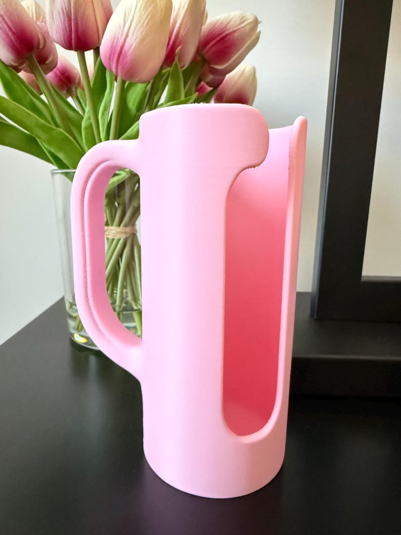 May include: A light pink cylindrical sleeve featuring a vertical cutout and a side handle. The object has a smooth, matte finish and is designed to wrap around a container, providing a grip for holding while maintaining an open front section.