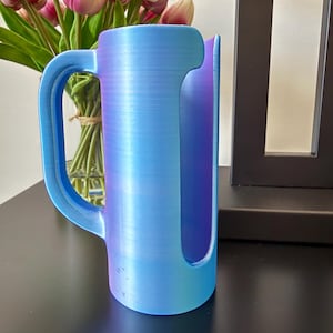 May include: A cylindrical mug-shaped holder with a vertical side handle and a large rectangular cutout on the front. The object features a smooth, iridescent finish that transitions between shades of light blue, purple, and magenta throughout.