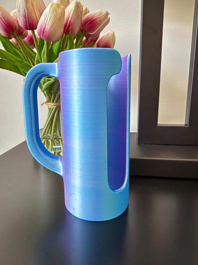 May include: A cylindrical mug-shaped holder with a vertical side handle and a large rectangular cutout on the front. The object features a smooth, iridescent finish that transitions between shades of light blue, purple, and magenta throughout.