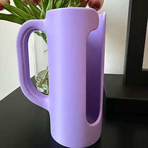 May include: A matte lavender-colored cylindrical holder featuring a large, curved handle on one side and a vertical, elongated cutout opening along the front face. The object has a smooth, uniform finish and a hollow interior structure.
