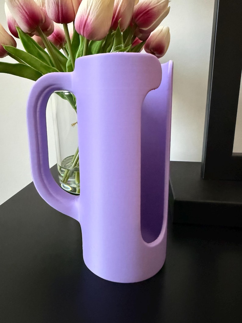 May include: A matte lavender-colored cylindrical holder featuring a large, curved handle on one side and a vertical, elongated cutout opening along the front face. The object has a smooth, uniform finish and a hollow interior structure.