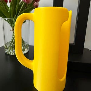 May include: A bright yellow plastic pitcher featuring a smooth, cylindrical body with a large, integrated handle on the side and a wide, open vertical cutout along the front edge, designed for pouring liquids with a simple, functional aesthetic.