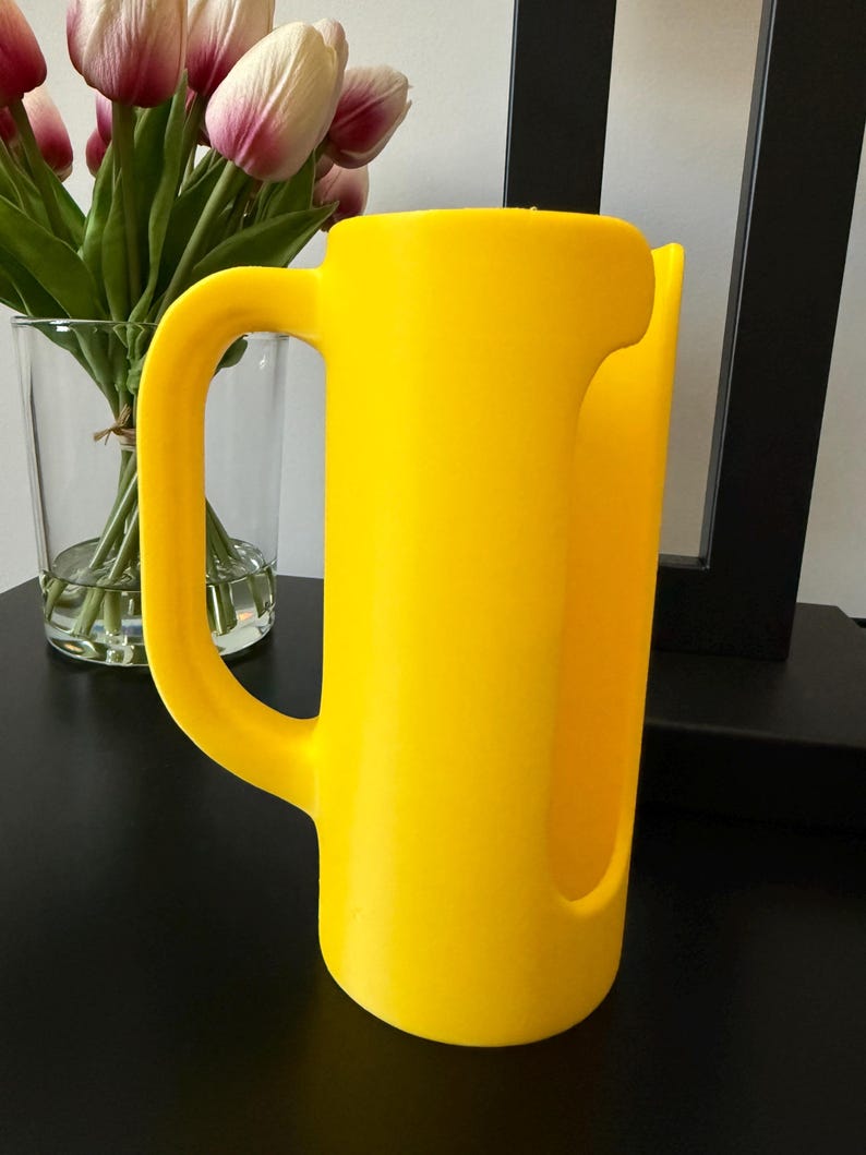 May include: A bright yellow plastic pitcher featuring a smooth, cylindrical body with a large, integrated handle on the side and a wide, open vertical cutout along the front edge, designed for pouring liquids with a simple, functional aesthetic.