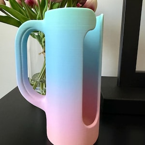 May include: A cylindrical cup holder with a handle, featuring a vertical gradient color transition from light blue at the top to a soft pink at the base. The object has a large, elongated vertical cutout on the side and a smooth, matte finish.
