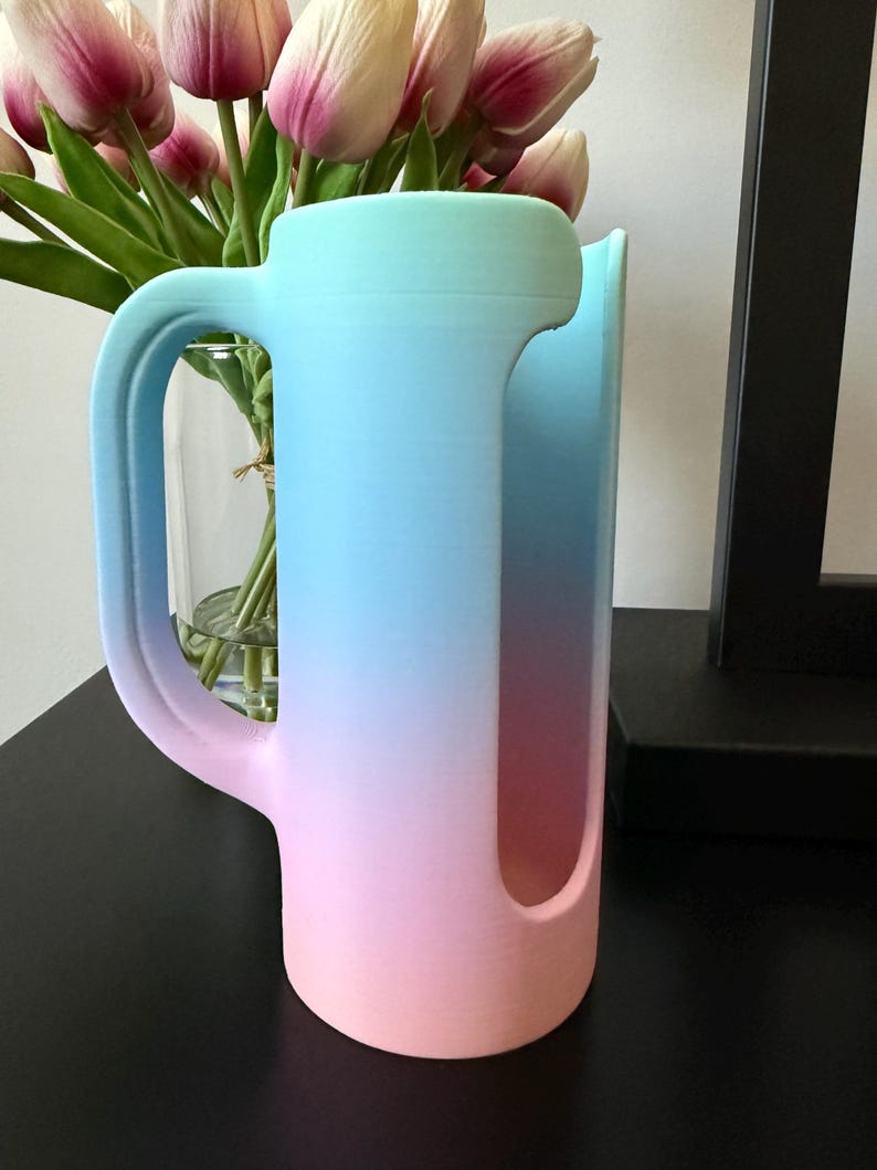 May include: A cylindrical cup holder with a handle, featuring a vertical gradient color transition from light blue at the top to a soft pink at the base. The object has a large, elongated vertical cutout on the side and a smooth, matte finish.