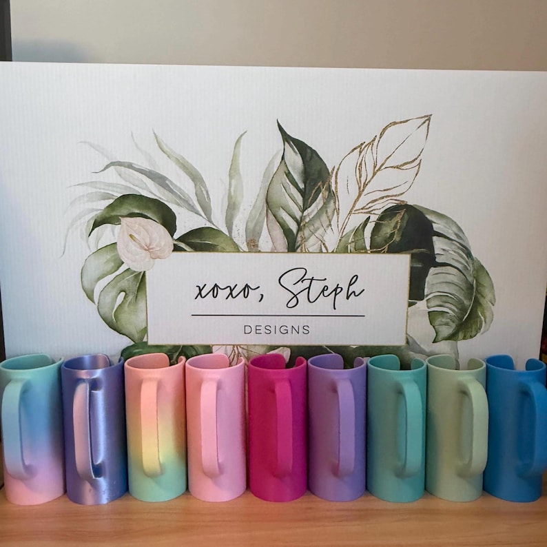 May include: A row of nine cylindrical mug wraps in various solid and gradient colors including blue, purple, pink, and teal, positioned in front of a white sign featuring green botanical illustrations and the text xoxo, Steph DESIGNS.